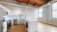 Located at 425 W Lakeside Ave in Cleveland's vibrant Lakeside neighborhood, this one-bedroom, one-bathroom loft-style unit showcases an impressive blend of industrial character and modern updates. The open-concept kitchen and living area is defined by striking exposed wood beam ceilings with track lighting, lending warmth and architectural interest to the space. The kitchen is thoughtfully appointed with white shaker cabinetry, a subway tile backsplash, quartz countertops, stainless steel appliances, and a peninsula that doubles as a breakfast bar. An in-unit washer is neatly integrated into the kitchen layout for added convenience. Light hardwood flooring runs throughout, enhancing the bright, airy feel of the unit. A hallway connects the main living area to the bedroom and bathroom, with modern wall sconces providing accent lighting along the way. The bedroom offers generous proportions, featuring whitewashed exposed brick walls and large windows that frame views of the Cleveland skyline and surrounding cityscape, filling the room with natural light. Two closets provide ample storage. The bathroom includes a full tub-shower combination with clean, white finishes. This unit masterfully combines historic industrial elements with contemporary design for a distinctive urban living experience.