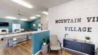 The Mountain View Village Clubhouse at 635 Northern Lights Blvd in Box Elder, South Dakota, is a thoughtfully designed single-floor community space that serves as a central hub for residents. The interior is defined by a bold teal accent color adorning key walls, which pairs elegantly with crisp white finishes, vaulted ceilings, and durable wood-look flooring throughout.

The main gathering area is generously sized and flooded with natural light from large windows dressed with patterned curtains, offering views of the surrounding community. This lounge features comfortable seating arrangements, a wall-mounted flat-screen TV, and a sleek white fireplace, creating a warm and inviting atmosphere ideal for socializing or relaxing.

A dedicated kitchenette area is equipped with white shaker-style cabinetry, a stainless steel refrigerator, microwave, and sink — providing convenient amenities for events and gatherings. Adjacent to the lounge, a pool table adds a recreational focal point to the space.

The entry area features a textured white shiplap accent wall displaying the Mountain View Village branding, complete with a seating nook and a navy storage cabinet, setting a polished first impression. Ceiling fans, recessed lighting, and ample ventilation round out the practical and comfortable design of this well-appointed clubhouse.