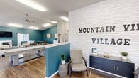 The Mountain View Village Clubhouse at 635 Northern Lights Blvd in Box Elder, South Dakota, is a thoughtfully designed single-floor community space that serves as a central hub for residents. The interior is defined by a bold teal accent color adorning key walls, which pairs elegantly with crisp white finishes, vaulted ceilings, and durable wood-look flooring throughout.

The main gathering area is generously sized and flooded with natural light from large windows dressed with patterned curtains, offering views of the surrounding community. This lounge features comfortable seating arrangements, a wall-mounted flat-screen TV, and a sleek white fireplace, creating a warm and inviting atmosphere ideal for socializing or relaxing.

A dedicated kitchenette area is equipped with white shaker-style cabinetry, a stainless steel refrigerator, microwave, and sink — providing convenient amenities for events and gatherings. Adjacent to the lounge, a pool table adds a recreational focal point to the space.

The entry area features a textured white shiplap accent wall displaying the Mountain View Village branding, complete with a seating nook and a navy storage cabinet, setting a polished first impression. Ceiling fans, recessed lighting, and ample ventilation round out the practical and comfortable design of this well-appointed clubhouse.