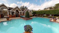 Located at 7827 Jefferson Place Circle in Baton Rouge, Louisiana, this single-story community facility showcases a well-appointed outdoor amenity area centered around a generously sized swimming pool with clear, turquoise water. The pool deck is finished with classic brick detailing and features a decorative island planter filled with colorful seasonal flowers, adding a vibrant focal point to the aquatic space. Lounge chairs are positioned poolside, offering comfortable spots for relaxation and sunbathing.

The adjacent clubhouse building is designed in a traditional Southern architectural style, featuring pitched rooflines, arched windows with decorative trim, white columns, and a combination of taupe siding and brick accents. A covered porch area extends from the clubhouse toward the pool, furnished with teal outdoor seating that creates a comfortable gathering space. Neatly trimmed topiary shrubs and manicured hedgerows frame the perimeter of the pool area, contributing to a polished and well-maintained landscape. Mature trees in the background provide natural shade and a lush, green backdrop. The overall ambiance is welcoming and resort-like, reflecting a thoughtfully designed outdoor environment suited for community leisure and social activity.