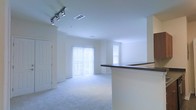 This single-floor residence offers a clean, open-concept layout designed to maximize both space and functionality. The living area is anchored by plush wall-to-wall carpeting that adds warmth and comfort underfoot, while crisp white walls create a bright, airy atmosphere throughout. Large sliding glass doors dressed with vertical blinds flood the main living space with an abundance of natural light, complemented by an additional side window that further enhances the sense of openness. A stylish track lighting fixture mounted on the ceiling provides adjustable task and ambient lighting, offering both practicality and a modern aesthetic touch. The kitchen area features a raised breakfast bar counter with a dark countertop surface, providing a natural division between the kitchen and living spaces while maintaining the open feel of the floor plan. Crown molding details along the ceiling add a refined architectural element to the interior. The layout flows seamlessly, making the space feel welcoming and well-proportioned. Ceiling vents are integrated discreetly throughout, ensuring efficient climate control. Overall, the space presents a light-filled, move-in-ready interior with thoughtful design details suited for comfortable everyday living.