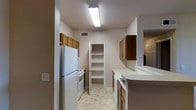 This single-story space includes a galley-style kitchen designed for efficient use of space. The kitchen is equipped with essential appliances, including a full-size refrigerator and a range, both finished in white. Medium-toned wood cabinetry lines both sides of the kitchen, providing generous storage both above and below the counter. The white countertops offer a clean, neutral workspace throughout. A built-in pantry with open shelving sits at the end of the kitchen corridor, adding further storage and organizational flexibility. The flooring features light-toned vinyl tile in a subtle pattern, contributing to the bright, clean aesthetic of the space. Overhead fluorescent lighting ensures the kitchen is well-lit. A cutout or pass-through on one side of the kitchen opens toward an adjacent area, enhancing the sense of connectivity within the layout. Ventilation is provided through a ceiling vent, and neutral wall tones throughout keep the space feeling open and cohesive. The overall design prioritizes practicality and functionality within a straightforward, single-level floor plan.