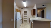 This single-story space includes a galley-style kitchen designed for efficient use of space. The kitchen is equipped with essential appliances, including a full-size refrigerator and a range, both finished in white. Medium-toned wood cabinetry lines both sides of the kitchen, providing generous storage both above and below the counter. The white countertops offer a clean, neutral workspace throughout. A built-in pantry with open shelving sits at the end of the kitchen corridor, adding further storage and organizational flexibility. The flooring features light-toned vinyl tile in a subtle pattern, contributing to the bright, clean aesthetic of the space. Overhead fluorescent lighting ensures the kitchen is well-lit. A cutout or pass-through on one side of the kitchen opens toward an adjacent area, enhancing the sense of connectivity within the layout. Ventilation is provided through a ceiling vent, and neutral wall tones throughout keep the space feeling open and cohesive. The overall design prioritizes practicality and functionality within a straightforward, single-level floor plan.