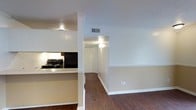 This single-floor space features an open-concept layout that connects the kitchen and living areas seamlessly. The kitchen is equipped with white upper cabinetry, a black electric range, and dark countertops, with a half-wall peninsula that defines the space without fully enclosing it. A structural column adds architectural character at the kitchen's edge. The flooring throughout is dark hardwood, providing a warm contrast to the neutral, light-toned walls. A textured ceiling runs through the main areas, adding subtle visual interest overhead. A hallway extending from the main living area leads to additional rooms, featuring a louvered bi-fold closet door and wall-mounted light fixture. HVAC vents are positioned in the ceiling for efficient climate control. The living area offers clean wall space, white baseboards, and a neutral palette that creates a versatile backdrop. Overall, the space maintains a simple, functional design with consistent finishes throughout, suited for everyday living.