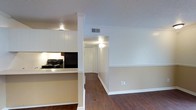 This single-floor space features an open-concept layout that connects the kitchen and living areas seamlessly. The kitchen is equipped with white upper cabinetry, a black electric range, and dark countertops, with a half-wall peninsula that defines the space without fully enclosing it. A structural column adds architectural character at the kitchen's edge. The flooring throughout is dark hardwood, providing a warm contrast to the neutral, light-toned walls. A textured ceiling runs through the main areas, adding subtle visual interest overhead. A hallway extending from the main living area leads to additional rooms, featuring a louvered bi-fold closet door and wall-mounted light fixture. HVAC vents are positioned in the ceiling for efficient climate control. The living area offers clean wall space, white baseboards, and a neutral palette that creates a versatile backdrop. Overall, the space maintains a simple, functional design with consistent finishes throughout, suited for everyday living.