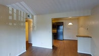 This single-floor residence offers a practical and well-connected layout designed for comfortable everyday living. The main living area is an open, airy space featuring warm hardwood flooring that extends seamlessly throughout, lending a sense of continuity and warmth. Neutral white walls provide a clean, versatile backdrop, while a textured ceiling adds a classic residential character.

The kitchen is directly accessible from the living area and is equipped with dark-toned cabinetry that provides generous storage, a black refrigerator, and countertop workspace. A pendant light fixture hangs above, adding a focused point of illumination that complements the functional design of the space. The kitchen layout is efficient and well-organized, making it easy to navigate.

Adjacent to the living area, a hallway leads to additional rooms, which are finished with the same consistent hardwood flooring and warm ambient lighting. A visible HVAC vent in the ceiling ensures climate control throughout the space.

Overall, the residence presents a cohesive, move-in-ready interior with a neutral color palette, dark wood cabinetry accents, and durable flooring — creating a balanced and practical living environment across its single floor.