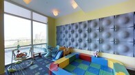 This cheerful children's playroom is thoughtfully designed to provide a safe and stimulating environment for young ones. The space is anchored by a striking feature wall covered in large, tactile upholstered panels in a deep grey tone, offering both visual interest and practical cushioning. Bright yellow walls complement the playful atmosphere, while recessed and flush-mount ceiling lights ensure even, welcoming illumination throughout.

A colorful plastic slide and a ride-on toy car serve as focal points within the play area, surrounded by a vibrant arrangement of modular foam play mats and soft climbing structures in primary colors — ideal for active, imaginative play. A small table and chairs set in a contrasting blue hue occupies one corner, providing a dedicated space for quieter activities such as drawing or crafts.

The room benefits from generous floor-to-ceiling windows along one wall, flooding the space with natural light and offering expansive urban skyline views. The patterned blue area rug grounds the room and adds another layer of color and texture. Overall, the space balances energetic design with child-safe materials, creating an engaging and functional play environment.