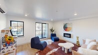 This thoughtfully designed living space offers a compact yet functional single-floor layout centered around a bright and inviting living room. The interior features clean white walls, smooth ceilings with recessed lighting, and warm light wood flooring that flows throughout. A sleek wall-mounted electric fireplace serves as a focal point on one wall, complemented by a vibrant circular abstract artwork that adds a bold splash of color to the space.

The living area is furnished with sculptural navy blue lounge chairs, a blush sofa, and an organically shaped white coffee table, all anchored by a rich brown cowhide-style area rug that adds texture and warmth. A classic arcade cabinet and a sun-motif wall print introduce a playful, eclectic character to the space, while a split-unit HVAC system ensures year-round comfort.

Large sliding glass doors flood the interior with natural light and provide direct access to a covered outdoor balcony. The balcony features light wood-toned flooring, navy railings, and seating areas that overlook lush tree canopy on one side and urban surroundings on the other — offering a versatile outdoor retreat. A mini split system and a dedicated hallway corridor contribute to the space's practical, well-organized layout. Overall, this living space strikes a balance between modern design and relaxed, creative living.