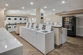 Located at 5600 Orangethorpe Ave in La Palma, California, this well-appointed shared laundry facility is designed to accommodate high-demand use with efficiency and comfort in mind. The single-floor space features a generous open layout illuminated by recessed lighting throughout, creating a bright and clean environment for residents.

Multiple top-loading washing machines are arranged in rows along a central island and perimeter walls, complemented by a bank of stacked dryers positioned along the back wall. Each machine is numbered for easy identification, and the modern units are equipped with digital displays for straightforward operation.

Granite-topped folding counters run along one side of the space, providing ample room for sorting and folding laundry. The wood-look laminate flooring adds a warm, residential feel to the utilitarian space, while crown molding along the ceiling lends a polished, finished appearance.

A snack and beverage vending machine is conveniently located near the entrance, adding a practical amenity for users during longer wash cycles. A sliding glass door at one end of the room allows natural light to filter in and provides access to an outdoor area. Safety features, including a fire extinguisher and clearly posted no-smoking signage, are present throughout. The overall ambiance is tidy, functional, and well-maintained.