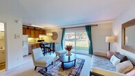 This efficiently designed single-floor unit offers a thoughtfully arranged open-concept layout that maximizes every inch of its footprint. The main living area is bright and welcoming, anchored by a large sliding glass door framed with teal curtains that floods the space with natural light and provides pleasant views of the surrounding greenery. Plush carpeted flooring runs throughout, adding warmth and comfort underfoot.

The living area flows seamlessly into the kitchen, which features warm honey-toned wood cabinetry, a full-size refrigerator, and a range, offering functional everyday cooking space. A cozy dining nook sits at the boundary between the kitchen and living area, making it an ideal spot for casual meals. A built-in closet with organizational shelving and hanging space provides ample storage, while a wall-mounted mirror and soft ambient lighting enhance the sense of openness.

The bathroom is finished with classic white tile walls and distinctive retro-style yellow fixtures, including a soaking tub with a decorative shower curtain, a pedestal-style vanity with under-sink cabinetry, and a medicine cabinet mirror. A dark open shelving unit adds a stylish contrast while offering additional storage. The patterned tile flooring completes the room's vintage-inspired character, giving the space a unique and memorable aesthetic.