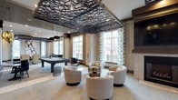Fifth Street Place is a thoughtfully designed single-floor communal space that blends contemporary aesthetics with comfortable, functional amenities. The open layout is anchored by a striking decorative ceiling panel featuring an intricate geometric lattice pattern in dark metal, which serves as a dramatic focal point overhead. Pendant lighting in geometric gold-toned frames complements the design, adding warmth and sophistication throughout the space.

A central lounge area is furnished with plush swivel chairs arranged around a low coffee table, creating an inviting setting for casual conversation or relaxation. Nearby, a full-size billiards table provides entertainment for guests and residents alike. A classic fireplace with a herringbone-patterned surround anchors one wall, paired with a large wall-mounted television, making it a natural gathering spot.

The dining and bar area features a generous granite-topped counter with rustic wooden barstools, offering a relaxed space for meals or socializing. Large windows dressed with patterned curtains allow ample natural light to filter in, with glass doors providing direct access to an outdoor area. The neutral color palette of the carpeting and tile flooring, combined with light brick wall accents and dark wood trim details, creates a cohesive and welcoming ambiance throughout.
