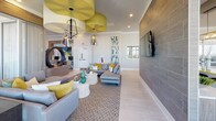 This thoughtfully designed amenity space offers a dynamic blend of functionality and contemporary style across a single floor. Floor-to-ceiling windows flood the interior with abundant natural light and frame sweeping elevated views of the surrounding cityscape, creating a bright and airy atmosphere throughout.

The lounge area serves as the heart of the space, anchored by a large textured tile accent wall with a wall-mounted flat-screen TV, plush sectional sofas, and striking pendant lighting featuring bold gold ceiling details. Adjacent to the lounge, a vibrant social area features a lime-green communal table with bar seating, decorative tiled wall panels, and intimate booth-style nooks ideal for casual gatherings.

A dedicated business center provides dual workstations recessed into a warm wood-paneled alcove, accompanied by comfortable lounge chairs and a coffee table — perfect for focused work or informal meetings. A private conference room with a sleek whitewashed table, upholstered chairs, and glass partition walls offers a more formal meeting environment.

The entertainment zone rounds out the space with a foosball table, shuffleboard, and direct access to an outdoor area, catering to recreational activities. Throughout, the design palette of soft grays, warm taupes, and pops of chartreuse and teal creates a cohesive, modern aesthetic that is both welcoming and visually engaging.