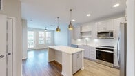 This well-designed single-floor residence offers a bright, open-concept layout that seamlessly connects the kitchen and living areas. The kitchen is a standout feature, showcasing white shaker-style cabinetry, a classic subway tile backsplash, and a full suite of stainless steel appliances including a French door refrigerator, electric range, built-in microwave, and dishwasher. A generously sized center island with a crisp white countertop provides additional prep space and serves as a natural gathering point. Metallic pendant lights add a contemporary touch overhead, while recessed lighting ensures the space remains bright throughout. Light wood-tone flooring flows throughout the main living areas, complementing the neutral color palette. The living area benefits from large windows and a glass-panel door that open to an outdoor balcony, inviting abundant natural light and views of the surrounding neighborhood. A ceiling fan enhances comfort in both the living space and the bedrooms. The bedrooms feature plush carpeting, ceiling fans, and windows with exterior views. The bathroom is clean and functional, outfitted with a tub-shower combination, subway tile surround, brushed nickel fixtures, and wood-tone flooring, maintaining the cohesive modern aesthetic found throughout the home.