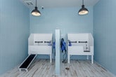 This single-room pet grooming and washing facility is thoughtfully designed with both function and comfort in mind. The space is defined by its soft powder-blue tile walls, which extend floor to ceiling in the wet zone, creating a clean, hygienic environment that is easy to maintain. Light wood-look plank flooring spans the open area, offering a warm contrast to the cool-toned walls and lending a welcoming feel to the space.

At the heart of the room is a built-in white dog wash station, complete with a gentle access ramp to assist pets of all sizes. A professional-grade blue pet dryer is wall-mounted directly beside the tub for convenient post-bath use, along with additional grooming accessories within easy reach. Industrial-style pendant lights hang from the ceiling, providing focused, warm illumination throughout the space.

A large window fitted with white blinds and a glass-panel exterior door allow generous natural light to fill the room while offering views of the surrounding outdoor area. A thermostat and a wall-mounted fire extinguisher cabinet reflect the practical safety considerations built into the design. Whimsical pet-themed wall art adds a playful touch, reinforcing the space's dedicated purpose. Overall, the facility strikes a balance between professional utility and an inviting, cheerful atmosphere.