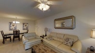 This single-floor space centers around a well-appointed living room designed for comfort and functionality. The room features neutral-toned walls that create a calm, inviting atmosphere, complemented by light-colored tile flooring partially covered by a patterned area rug that adds texture and warmth underfoot.

The seating arrangement includes a full-sized leather sofa and a matching leather armchair, both in a soft cream finish, providing ample seating while maintaining a cohesive aesthetic. Decorative patterned throw pillows accent the sofa, adding visual interest to the understated color palette.

A ceiling fan with an integrated light fixture is centrally mounted, offering both ambient lighting and air circulation. Additional lighting is provided by a classic table lamp positioned on an end table between the seating pieces.

Wall décor includes a large ornate-framed mirror that reflects light and enhances the sense of space, as well as a framed artwork piece near the entryway. A compact workspace or dining area is partially visible in the background, suggesting a versatile use of the available space. The interior entry door is clearly visible, contributing to the practical, straightforward layout of this single-level home.