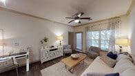This single-floor residence offers a thoughtfully designed layout that combines comfort and functionality. The living area serves as the heart of the home, featuring warm dark hardwood-style flooring, crown molding, and a ceiling fan that add a polished touch to the space. Large windows dressed with layered curtains and blinds allow natural light to fill the room, while a decorative wall mirror enhances the sense of openness. The living area flows seamlessly into the kitchen, which is equipped with dark cabinetry, black appliances including a refrigerator and range, and a peninsula countertop with light stone-style surfaces and bar stool seating. Pendant lighting and a track light system illuminate the kitchen, creating a warm and practical cooking environment. The bedroom is a serene retreat with plush carpeting, a ceiling fan, and a window that invites soft natural light. Neutral tones and clean lines throughout the room contribute to a calm, restful ambiance. The bathroom is accessed via a hallway and features dark vanity cabinetry, a large mirror, and dark hardwood-style flooring, maintaining the cohesive design language of the residence. Overall, the space presents a harmonious blend of contemporary style and everyday practicality.