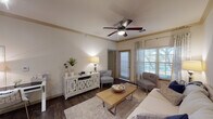 This single-floor residence offers a thoughtfully designed layout that combines comfort and functionality. The living area serves as the heart of the home, featuring warm dark hardwood-style flooring, crown molding, and a ceiling fan that add a polished touch to the space. Large windows dressed with layered curtains and blinds allow natural light to fill the room, while a decorative wall mirror enhances the sense of openness. The living area flows seamlessly into the kitchen, which is equipped with dark cabinetry, black appliances including a refrigerator and range, and a peninsula countertop with light stone-style surfaces and bar stool seating. Pendant lighting and a track light system illuminate the kitchen, creating a warm and practical cooking environment. The bedroom is a serene retreat with plush carpeting, a ceiling fan, and a window that invites soft natural light. Neutral tones and clean lines throughout the room contribute to a calm, restful ambiance. The bathroom is accessed via a hallway and features dark vanity cabinetry, a large mirror, and dark hardwood-style flooring, maintaining the cohesive design language of the residence. Overall, the space presents a harmonious blend of contemporary style and everyday practicality.