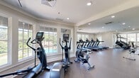 This well-appointed fitness center in Auburn, Alabama, offers a generously sized, single-floor layout designed to accommodate a comprehensive range of workout activities. The facility is organized into distinct zones, seamlessly connected through wide, open passageways that promote an airy, unobstructed flow throughout the space.

The cardio area is lined with large, multi-pane windows that flood the room with natural light and provide views of the surrounding outdoor pool and green spaces. Equipment includes multiple treadmills, elliptical trainers, and stationary bikes, all featuring integrated digital displays. Full-length wall mirrors amplify the sense of space and allow users to monitor their form.

The strength training section features a vaulted ceiling with ceiling fans, contributing to an open and comfortable atmosphere. Large windows continue to bring in abundant natural light. This area is equipped with a variety of selectorized weight machines, free-weight stations, barbells, and a full dumbbell rack. Floor-to-ceiling mirrors line the walls here as well.

Throughout, the facility features durable rubberized flooring, recessed lighting, and a neutral color palette of warm whites and soft grays. The clean architectural detailing, including crown molding and classic panel trim, lends a refined, upscale character to the functional fitness environment.