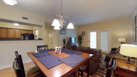 This single-floor space showcases an open-concept layout that seamlessly connects the dining area, living room, and kitchen. The dining area is anchored by a warm-toned wooden table set for multiple guests, accented by a brushed nickel multi-light chandelier that serves as a focal point overhead. Dark wood chairs with slatted backs complement the table's rich finish, while blue striped placemats add a touch of color.

Adjacent to the dining space, the living area features hardwood flooring, a comfortable upholstered sofa, and a media console with a flat-screen television. A patterned area rug defines the seating zone, adding texture and warmth to the space. A glass-paned exterior door allows natural light to filter in, brightening the interior and offering a connection to the outdoors.

To the side, a pass-through opening provides a glimpse into the kitchen, suggesting convenient flow between spaces for everyday living and entertaining. Neutral-toned walls and classic trim throughout the space contribute to a cohesive and welcoming aesthetic that balances practicality with comfort.