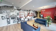 This single-floor space makes a strong first impression with its dynamic, urban-inspired design. The centerpiece of the lobby is a large-scale, graffiti-style mural that spans an entire accent wall, featuring bold red lettering spelling "ELAAN" alongside black-and-white cityscape illustrations, street art motifs, and typographic elements that evoke a vibrant metropolitan aesthetic. The mural creates a dramatic visual focal point that sets the tone for the entire space.

The seating area is thoughtfully arranged with contemporary blue sofas and a glass-topped coffee table with a warm wood base, offering a comfortable and stylish gathering space. Light hardwood flooring runs throughout, providing a clean contrast to the expressive wall art above. Recessed ceiling lighting ensures the area is well-lit and functional.

A bold red accent wall is visible to the right, reinforcing the space's strong color palette of red, white, black, and blue. Additional amenities, including what appears to be a beverage station, are integrated into the layout, enhancing the usability of the communal area. Overall, the space balances artistic expression with practical, welcoming design.