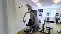 This single-floor fitness center is designed to support a comprehensive workout experience with a thoughtful selection of exercise equipment. At the forefront of the space stands a Life Fitness multi-station strength machine, offering a wide range of resistance training options including lat pulldown, chest press, and cable attachments — making it suitable for full-body strength training. The room is finished with neutral-toned walls and carpet flooring, contributing to a clean and functional environment. Natural light filters in through a window, while recessed ceiling lighting ensures the space remains well-lit throughout the day. A wall-mounted flat-screen television is positioned for easy viewing during workouts, adding a layer of comfort and entertainment. Toward the rear of the space, additional cardio equipment — including what appears to be a treadmill and an elliptical machine — is visible, further expanding the range of available fitness options. The mirrors along the wall enhance the sense of openness while allowing users to monitor their form during exercise. Overall, the fitness center is efficiently organized and well-maintained, providing a practical and inviting environment for a variety of workout routines.