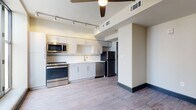 This single-floor unit offers a contemporary design with a practical and open layout. The main living area is bright and airy, benefiting from multiple large, black-framed windows that allow abundant natural light to fill the space. A glass door provides direct access to an outdoor balcony, offering open sky views. A ceiling fan overhead adds both function and a modern touch to the living space.

The kitchen is well-appointed and efficiently designed, featuring stainless steel appliances including a range, microwave, dishwasher, and refrigerator. White cabinetry with clean-lined hardware lines the walls, complemented by a white subway tile backsplash, light countertops, and a double stainless steel sink. Track lighting overhead ensures the kitchen is well-lit.

Wood-style plank flooring extends consistently throughout the living and kitchen areas, providing a warm yet modern aesthetic. Dark baseboards offer a sharp contrast against the neutral walls.

The bathroom is clean and straightforward, featuring a full tub-shower combination with white tile surround. Overall, the unit maintains a cohesive, modern aesthetic with neutral tones throughout.