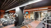 This fitness center is designed to accommodate a comprehensive workout experience across a single level. The space features an industrial aesthetic, highlighted by an exposed ceiling with visible ductwork and structural elements finished in dark tones, lending the gym a modern, urban feel. Bright LED strip lighting runs along the perimeter, ensuring the entire floor is well-illuminated throughout.

The gym floor is covered in durable rubber flooring in a distinctive two-tone pattern, providing both safety and comfort during exercise. Bold orange accent walls energize the environment and contrast effectively with the dark structural columns that anchor the space.

The cardio section is stocked with a row of treadmills and elliptical-style machines, while the strength training area includes a multi-function cable and weight machine system suitable for a variety of resistance exercises. A mounted flat-screen monitor is positioned centrally, offering entertainment or instructional content for users during workouts.

The layout is thoughtfully organized to allow users to move freely between equipment zones without congestion. The combination of industrial design, vibrant color choices, and a diverse selection of fitness equipment makes this a functional and motivating environment suited for both cardio and strength training routines.