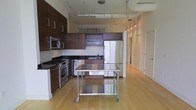 This single-floor space showcases a contemporary open-plan layout that seamlessly blends functionality with modern design. The kitchen is a standout feature, equipped with rich dark wood cabinetry that contrasts elegantly against dark granite countertops. Full stainless steel appliances, including a gas range, built-in microwave, dishwasher, and a large refrigerator, provide a professional-grade cooking environment. A mobile stainless steel prep table on casters sits at the center, offering flexible workspace and an industrial touch. Track lighting runs along the ceiling, casting adjustable illumination across the kitchen and into the adjoining open area, which is further brightened by pendant lights. Warm-toned hardwood flooring extends throughout the space, lending a sense of continuity and warmth that complements the modern palette. The open floor plan creates a generous, airy feel, with the kitchen flowing naturally into the living and corridor areas. Light-toned walls and high ceilings amplify the brightness, making the entire space feel expansive and welcoming. The overall ambiance is clean, modern, and well-appointed, suited for both everyday living and entertaining.