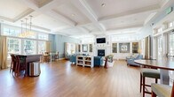 Located at 299 Colt Highway in Farmington, Connecticut, this single-floor community lounge is a thoughtfully designed gathering space that blends classic New England architecture with comfortable, modern furnishings. The room immediately impresses with its soaring coffered ceilings trimmed in crisp white molding, which add architectural depth and a sense of grandeur throughout the open-plan layout.

Warm hardwood flooring extends across the entire space, complementing the soft blue-gray walls accented by white wainscoting. A central fireplace flanked by white built-in shelving and cabinetry serves as the focal point of the main seating area, where plush blue sectional sofas and accent chairs invite relaxation. Adjacent built-in shelving units provide additional display and storage space.

A dedicated kitchenette area features rich dark wood cabinetry, granite countertops, a wall-mounted television, and a built-in bar-height island with seating — ideal for casual dining or socializing. Large multi-pane windows flood the space with abundant natural light and offer views of the surrounding greenery and outdoor amenities. French doors along one wall open directly to an outdoor patio or terrace, seamlessly connecting the interior to the exterior environment. The overall ambiance is bright, welcoming, and refined.