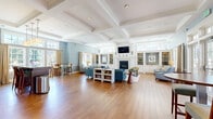 Located at 299 Colt Highway in Farmington, Connecticut, this single-floor community lounge is a thoughtfully designed gathering space that blends classic New England architecture with comfortable, modern furnishings. The room immediately impresses with its soaring coffered ceilings trimmed in crisp white molding, which add architectural depth and a sense of grandeur throughout the open-plan layout.

Warm hardwood flooring extends across the entire space, complementing the soft blue-gray walls accented by white wainscoting. A central fireplace flanked by white built-in shelving and cabinetry serves as the focal point of the main seating area, where plush blue sectional sofas and accent chairs invite relaxation. Adjacent built-in shelving units provide additional display and storage space.

A dedicated kitchenette area features rich dark wood cabinetry, granite countertops, a wall-mounted television, and a built-in bar-height island with seating — ideal for casual dining or socializing. Large multi-pane windows flood the space with abundant natural light and offer views of the surrounding greenery and outdoor amenities. French doors along one wall open directly to an outdoor patio or terrace, seamlessly connecting the interior to the exterior environment. The overall ambiance is bright, welcoming, and refined.