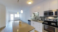 This single-floor residence offers a thoughtfully designed open-concept layout that seamlessly connects the kitchen and living areas. The kitchen is a standout feature, showcasing crisp white cabinetry, a neutral-toned countertop, and a striking subway tile backsplash in shades of grey that adds a contemporary flair. Stainless steel appliances, including a microwave and dishwasher, provide both functionality and a polished aesthetic. A double-basin sink with a modern arc faucet sits beneath under-cabinet lighting, enhancing workspace visibility. A breakfast bar-style peninsula extends the counter space, making it ideal for casual dining or entertaining. Pendant lighting hangs elegantly above, complementing the flush-mount ceiling fixture overhead. The open living area beyond the kitchen benefits from large windows that draw in generous amounts of natural light, creating an airy and welcoming atmosphere throughout. The flooring transitions smoothly between spaces, tying the overall design together with a clean, cohesive look. The combination of neutral tones, modern fixtures, and functional design elements makes this a well-appointed and versatile living space.