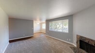 Located at 9900 SW Conestoga Dr in Beaverton, Oregon, this single-floor, 2-bedroom, 1-bathroom home offers a well-organized layout across a comfortable footprint. The living room is a standout space, featuring plush carpeting, neutral gray walls, a textured ceiling, and a striking stone-surround fireplace that serves as a natural focal point. Large windows with blinds fill the room with natural light and offer views of mature trees outside. The kitchen is appointed with white shaker-style cabinetry, dark granite-look countertops, a peninsula for added workspace, and warm wood-look flooring that extends into the dining area. A ceiling fan overhead adds comfort to the dining space. The hallway connects the two carpeted bedrooms—including a spacious primary bedroom—and the full bathroom. The bathroom features a white vanity with dark countertops, a large mirror, and wood-look flooring. Three closets throughout the home provide ample storage. Clean gray walls and white trim carry a consistent, modern aesthetic throughout. The entrance and hallway transition smoothly between all spaces, making the layout feel cohesive and functional.