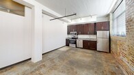 This urban-industrial unit at 144 4th St in Burlington, NC offers a distinctive loft-style aesthetic across a single floor. Polished concrete floors and exposed metal ductwork run throughout, complemented by white-painted wood plank ceilings with ceiling fans. The kitchen features dark espresso cabinetry, granite countertops, and a full suite of stainless steel appliances, including a range, microwave, dishwasher, and refrigerator, with track lighting overhead. A striking exposed brick accent wall with large windows floods the living area with natural light. Bedrooms include closets with built-in shelving, and the bathrooms are appointed with granite vanities, dark cabinetry, and full tub-shower combinations. Clerestory windows in select rooms add additional light while maintaining privacy.