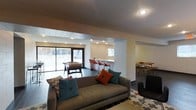 This single-floor space is thoughtfully designed for comfort and social engagement, offering a variety of distinct zones that flow seamlessly into one another. The central lounge area anchors the space with a plush, contemporary sofa adorned with colorful teal and orange accent pillows, set atop a patterned area rug that adds a playful touch to the modern aesthetic. Recessed lighting throughout provides a warm, inviting ambiance across the open layout. Adjacent to the lounge, a game area features a foosball table positioned near large sliding glass doors that flood the space with natural light and offer views to an outdoor area. A built-in bar-style counter with vibrant orange bar stools serves as a casual gathering spot, illuminated by track lighting that highlights the clean, white surfaces. A dedicated dining area with a wooden table and coordinating chairs provides a more structured setting for shared meals or group activities. The combination of concrete block accents, tile flooring, and modern furnishings gives the space a contemporary, energetic character well-suited for community use. The open sightlines between zones encourage interaction while still allowing each area to serve its distinct purpose.