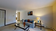 This single-floor residence offers a thoughtfully designed layout that combines comfort and style. The living room serves as the heart of the space, featuring soft, neutral wall tones and plush wall-to-wall carpeting that create a warm and inviting atmosphere. A classic sofa dressed with vibrant blue and brown accent pillows anchors the seating area, complemented by a glass-top coffee table and a coordinating side table that add a refined touch. A stylish table lamp provides warm ambient lighting, enhancing the room's cozy character. A colorful piece of wall art depicting a charming architectural scene adds a focal point and a pop of color to the otherwise understated palette. The smooth, white ceiling contributes to a sense of openness throughout the room. An open passageway connects the living area to adjacent spaces, including what appears to be a kitchen or dining area, suggesting a practical and connected floor plan. The overall ambiance is calm and composed, with décor choices that balance casual comfort with tasteful elegance, making it a welcoming environment for everyday living.