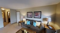 This single-floor space features a living room designed for comfort and casual everyday living. The room is anchored by a plush, grey upholstered sofa dressed with a mix of cream and teal accent pillows, adding a pop of color against the neutral palette. A glass-top coffee table sits in front of the sofa, complementing the warm wood tones present throughout the room, including a classic side table topped with a table lamp that provides soft ambient lighting. The walls are painted in a muted sage-green tone, creating a calm and inviting atmosphere. Two matching framed wall art pieces featuring floral motifs are displayed above the sofa, adding a decorative focal point to the space. The flooring is carpeted in a light beige tone, contributing to the room's overall warmth. A white paneled entry door is visible to one side, providing a clean architectural detail. The layout is straightforward and functional, making efficient use of the available space while maintaining an open, uncluttered feel. Overall, the space presents a relaxed and comfortable environment suitable for everyday living.