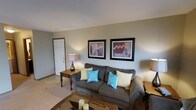 This single-floor space features a living room designed for comfort and casual everyday living. The room is anchored by a plush, grey upholstered sofa dressed with a mix of cream and teal accent pillows, adding a pop of color against the neutral palette. A glass-top coffee table sits in front of the sofa, complementing the warm wood tones present throughout the room, including a classic side table topped with a table lamp that provides soft ambient lighting. The walls are painted in a muted sage-green tone, creating a calm and inviting atmosphere. Two matching framed wall art pieces featuring floral motifs are displayed above the sofa, adding a decorative focal point to the space. The flooring is carpeted in a light beige tone, contributing to the room's overall warmth. A white paneled entry door is visible to one side, providing a clean architectural detail. The layout is straightforward and functional, making efficient use of the available space while maintaining an open, uncluttered feel. Overall, the space presents a relaxed and comfortable environment suitable for everyday living.