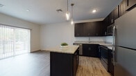This single-floor residence showcases a thoughtfully designed open layout that seamlessly connects the kitchen and living areas. The kitchen is a standout feature, equipped with dark espresso shaker-style cabinetry reaching toward the ceiling for ample storage, granite countertops, and a full suite of stainless steel appliances including a dishwasher, range, over-the-range microwave, and refrigerator. A central island with matching granite provides additional prep space and casual seating, while two pendant lights with glass jar-style fixtures add a stylish focal point overhead. Wood-look plank flooring runs throughout the main living areas, contributing to a clean, contemporary aesthetic.

The bedrooms are carpeted for comfort and finished in neutral tones, with at least one room featuring a walk-in closet and a window that brings in natural light. The bathroom maintains the design consistency of the rest of the unit, featuring a dark vanity with granite countertop and a tub-shower combination with a sliding glass rod.

A dedicated in-unit laundry area with a stacked washer and dryer is tucked into its own closet space, adding functional convenience. Vertical blinds on a sliding glass door allow for privacy and light control. The overall ambiance is modern, clean, and move-in ready.