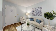 Located at 2631 Leaphart Rd in West Columbia, South Carolina, this thoughtfully designed single-floor suite offers a comfortable and functional living environment. The main living and sleeping area is an open, airy space finished with warm hardwood-style flooring and soft sage-green walls that create a calm, welcoming ambiance. A large sliding glass door with vertical blinds allows natural light to fill the room, while a cozy seating arrangement — including a loveseat, floral accent chairs, and a glass-top coffee table — defines a dedicated living area alongside the sleeping space. Tasteful wall art and greenery add warmth and personality throughout. The compact kitchen features wood-trimmed white cabinetry, a full-size refrigerator, an electric range, and a sink, providing all essential cooking amenities within an efficient footprint. Light wood-style flooring carries through the kitchen, contributing to a cohesive aesthetic. A small dining nook with a round table is conveniently positioned nearby. The bathroom is equipped with an accessibility-focused design, featuring a walk-in style tub with sturdy safety grab bars and a handheld showerhead, prioritizing ease of use and safety. Overall, the suite presents a clean, serene, and well-appointed environment well-suited for comfortable daily living.