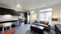 This single-floor residence showcases a seamlessly integrated open-concept layout that combines the kitchen and living area into one cohesive, well-appointed space. The kitchen is outfitted with dark espresso cabinetry that creates a striking contrast against light granite countertops and a full suite of stainless steel appliances, including a range, microwave, refrigerator, and dishwasher. Track lighting overhead ensures excellent task illumination throughout the cooking area. The living space flows naturally from the kitchen, anchored by a neutral color palette with warm accent tones. Large windows along the exterior wall flood the room with abundant natural light, creating an airy and inviting atmosphere. Light-toned hardwood-style flooring runs throughout, lending consistency and a sense of openness to the entire floor plan. The living area comfortably accommodates a sectional sofa and ottoman, making it ideal for relaxing or entertaining. The neutral walls and clean architectural lines contribute to a modern yet welcoming ambiance. Overall, this space offers a thoughtfully designed environment that balances style and practicality, with quality finishes and a bright, open feel throughout.