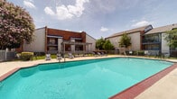 Located at 2929 Crossview Dr in Houston, Texas, this outdoor amenity area centers around a generously sized swimming pool with vivid turquoise water and a classic brick-edged surround. The pool deck is finished in light-colored concrete, providing ample space for lounging and circulation around the full perimeter of the pool. A freestanding pergola structure on one side of the deck offers a shaded gathering spot, accompanied by several modern lounge chairs positioned to take advantage of the open sky and Houston sunshine. Adjacent to the pool, a single-story clubhouse building features a distinctive blue accent canopy, vertical siding, and a glass-fronted facade that opens toward the pool area, suggesting indoor-outdoor functionality for gatherings or community use. The space is enclosed by a metal security fence with gated access, ensuring a safe and private environment. Mature trees and flowering shrubs, including a prominent blooming crape myrtle, add natural color and shade to the surroundings. The broader complex features multi-story residential buildings with balconies that overlook the pool courtyard, creating a cohesive community atmosphere. This well-appointed outdoor space serves as a central recreational hub within the property.