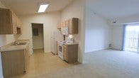 This single-floor space is defined by its vaulted ceilings, which lend a sense of openness and airiness throughout the interior. The kitchen area features light wood upper cabinetry providing ample storage, complemented by white appliances including a freestanding electric range, over-the-range microwave, and a full-size refrigerator. The countertop extends alongside the cabinetry, offering a functional workspace. The kitchen is finished with neutral-toned tile flooring that transitions seamlessly into a carpeted living area, creating a clear yet open delineation between the two zones. A skylight installed in the kitchen ceiling draws in natural light, brightening the space without relying solely on artificial lighting. The walls throughout are finished in a clean, neutral white, contributing to the bright and understated aesthetic. A hallway visible from the kitchen leads to additional areas of the unit, with what appears to be an electrical panel accessible nearby. The overall ambiance is clean, functional, and straightforward, with an open-concept layout that maximizes the sense of space within a compact single-floor footprint.