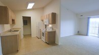 This single-floor space is defined by its vaulted ceilings, which lend a sense of openness and airiness throughout the interior. The kitchen area features light wood upper cabinetry providing ample storage, complemented by white appliances including a freestanding electric range, over-the-range microwave, and a full-size refrigerator. The countertop extends alongside the cabinetry, offering a functional workspace. The kitchen is finished with neutral-toned tile flooring that transitions seamlessly into a carpeted living area, creating a clear yet open delineation between the two zones. A skylight installed in the kitchen ceiling draws in natural light, brightening the space without relying solely on artificial lighting. The walls throughout are finished in a clean, neutral white, contributing to the bright and understated aesthetic. A hallway visible from the kitchen leads to additional areas of the unit, with what appears to be an electrical panel accessible nearby. The overall ambiance is clean, functional, and straightforward, with an open-concept layout that maximizes the sense of space within a compact single-floor footprint.