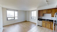 This single-floor space offers a clean, functional layout with an open-concept living and kitchen area that maximizes both light and usability. The living area features light-toned hardwood flooring that transitions seamlessly into tile near the kitchen, creating a natural visual separation between spaces. Multiple large windows flood the interior with natural light while framing elevated views of the surrounding cityscape and greenery, giving the space an airy, open feel.

The kitchen is positioned to the side of the main living area and is outfitted with warm honey-toned wood cabinetry, dark granite or stone countertops, and a stainless steel dishwasher. The contrast between the dark counters and warm wood tones lends the kitchen a classic, practical aesthetic.

The walls are finished in crisp white throughout, complementing the light flooring and contributing to a bright, neutral palette that suits a variety of interior styles. The ceiling features a smooth finish, further enhancing the sense of openness. Overall, the space presents a straightforward and well-lit layout, ideal for comfortable everyday living, with city views and natural light serving as its most notable highlights.