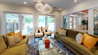 Located at 1100 Cannonball Run in Knightdale, NC, this single-floor community clubhouse is a thoughtfully designed gathering space that balances comfort and style. The lounge area serves as the heart of the space, featuring plush olive-green sofas accented with mustard and neutral throw pillows, arranged around a sleek glass-top coffee table. Striking white sculptural pendant chandeliers hang overhead, while large floor-to-ceiling windows and glass doors flood the room with abundant natural light and provide views of the surrounding greenery. A bold geometric accent wall and a shiplap-paneled wall with a wall-mounted flat-screen television add distinct character to the lounge. The adjacent bar-height dining area is defined by a vivid orange ceiling and warm pendant lighting above a long communal wood table surrounded by white crossback bar stools, all anchored by a rich blue patterned area rug. This area opens directly to an outdoor pool deck through glass double doors. The fully equipped kitchen features white shaker cabinetry with gold hardware, a blue mosaic tile backsplash, stone countertops, stainless steel appliances including a double wall oven and side-by-side refrigerator, open wood shelving, and light wood-look flooring throughout — making it ideal for hosting events and gatherings.