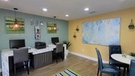 This single-floor space presents a thoughtfully designed interior that blends functionality with a warm, inviting aesthetic. The workspace is defined by a striking two-tone color scheme, featuring a muted teal accent wall paired with a soft golden-yellow wall, creating a balanced and energizing atmosphere. Recessed ceiling lighting provides consistent, even illumination throughout, complemented by a geometric pendant light fixture that adds a contemporary touch above the central work area.

The workstation area is anchored by a sleek counter featuring a marble-patterned surface atop dark cabinetry, providing ample workspace alongside a desktop computer and printer setup. Decorative diamond-shaped wall sconces add visual interest across both accent walls, while a large abstract canvas artwork dominates the yellow wall, serving as a bold focal point. A magazine rack and framed wall lettering contribute to the space's identity and character.

Wood-look flooring runs throughout, offering a clean, durable foundation that ties the room together. A striped area rug near the entrance adds a subtle layer of texture. A potted plant introduces a natural element, enhancing the overall freshness and approachability of this well-organized, single-level space.