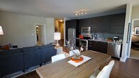 This single-floor space showcases an open-concept design that integrates the kitchen, dining, and living areas into one cohesive and functional layout. The kitchen is outfitted with dark espresso cabinetry extending to the upper walls, granite countertops, and stainless steel appliances including a refrigerator, microwave, and range. A subway tile backsplash and under-cabinet lighting add refined detail to the culinary space.

The dining area features a rustic wood plank table paired with white chairs, creating a warm contrast against the contemporary surroundings. A decorative centerpiece with a natural branch arrangement adds an artistic focal point to the dining space.

The living area is furnished with a plush dark grey sectional sofa, grounded by a patterned area rug. A table lamp with a white shade provides soft ambient lighting. The open sightlines between the living and dining areas create a sense of spaciousness and ease of flow.

A hallway visible from the main space leads to additional rooms, hinting at further private areas beyond. Neutral wall tones and consistent dark hardwood flooring tie the entire space together, giving it a cohesive, modern aesthetic.