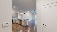 Located in Huntersville, NC, this well-appointed 2-bedroom, 2-bathroom single-floor residence offers a thoughtfully designed open-concept layout. The kitchen serves as a centerpiece of the home, showcasing gray shaker-style cabinetry with matte black hardware, dark quartz countertops, a subway tile backsplash, stainless steel appliances — including a refrigerator, range, and microwave — and a peninsula with an undermount sink and pendant lighting overhead. Hardwood-style flooring flows seamlessly from the kitchen into the living room, which features vaulted ceilings, a ceiling fan, and French doors that invite natural light and outdoor views. Both bedrooms are carpeted and equipped with ceiling fans; the primary bedroom is generously sized with its own closet. The two bathrooms are finished with dark quartz vanity tops, gray cabinetry, matte black fixtures, and wood-look flooring, with one bathroom offering a full tub-shower combination with white tile surround. A dedicated laundry room houses a full-size washer and dryer with overhead shelving. A connecting hallway ties all spaces together, completing this clean, modern, move-in-ready home.