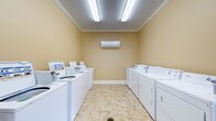 This communal laundry facility is designed for efficiency and comfort, offering a clean and organized environment for residents. The room is lined on both sides with a generous row of top-loading washing machines and matching dryers, providing ample capacity for multiple users. The warm beige walls are accented with crisp white crown molding, lending the space a polished, well-maintained appearance. Bright fluorescent ceiling fixtures illuminate the entire room evenly, ensuring excellent visibility at any time of day. A wall-mounted ductless air conditioning unit is centrally positioned to keep the space comfortable during use. The floor is finished with neutral, stone-patterned ceramic tiles that are both durable and easy to maintain. A detergent vending machine is conveniently located near the entrance, adding to the practical layout of the room. The space is thoughtfully arranged to allow easy movement between machines, making the laundry experience straightforward and accessible. Overall, this facility reflects a commitment to cleanliness, functionality, and resident convenience.