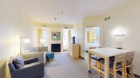 This thoughtfully designed hotel suite offers a comfortable and functional layout across a single floor, making it well-suited for extended stays or group travel. The living area serves as the heart of the suite, featuring a plush sectional sofa, a teal ottoman, a wall-mounted TV, and a cozy fireplace framed by colorful modern artwork. Natural light filters in through curtained windows, contributing to the suite's warm, inviting ambiance with soft yellow and cream tones throughout.

Adjacent to the living area is a dining space with a white-topped table and seating for four, flowing seamlessly into a fully equipped kitchen. The kitchen is outfitted with stainless steel appliances including a full-size refrigerator, oven, microwave, and dishwasher, along with ample cabinetry — ideal for preparing meals in-suite.

The suite includes two bedrooms, each furnished with a king-size bed, light wood headboard, coordinating nightstands, and soft blue accent walls that lend a calm, restful atmosphere. One bedroom features a wall-mounted TV and mirrored closet doors, while both offer natural light from exterior-facing windows.

Two bathrooms serve the suite: one featuring a clean vanity with a wide mirror and warm lighting, and the other with a full tub-shower combination with tile flooring — both finished in crisp white for a clean, polished look.